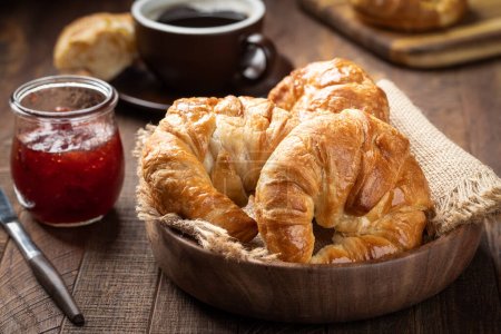 Photo for Fresh croissants in a wooden bowl with cup of coffee and jar of strawberry preserves in background - Royalty Free Image