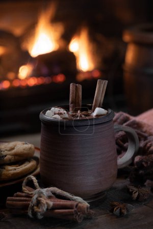 Photo for Mug of hot chocolate, marshmallows, anise and cinnamon sticks with blanket and plate of cookies on wooden table.  Fireplace burning in background - Royalty Free Image