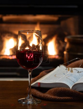 Photo for Glass of red wine with blanket and book on wooden table.  Burning fireplace in bqckground - Royalty Free Image