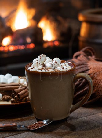 Photo for Cup of hot chocolate and marshmallows with blanket on wooden table.  Fireplace burning in background - Royalty Free Image