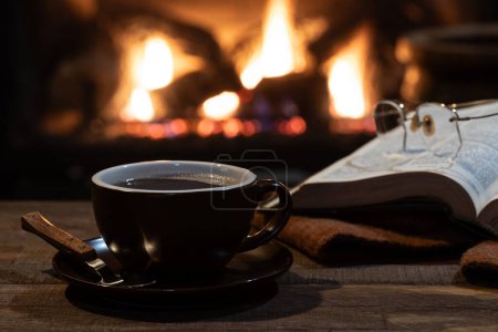 Photo for Cup of coffee with open book and blanket with a burning fireplace in background - Royalty Free Image