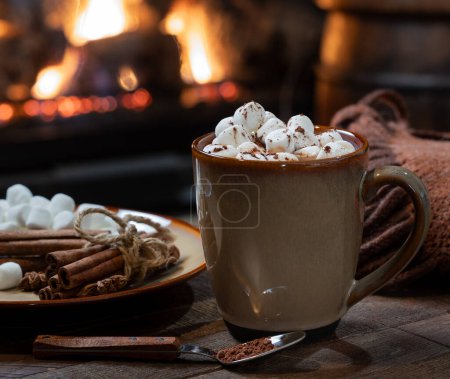 Foto de Copa de chocolate caliente y malvaviscos con manta sobre mesa de madera. Chimenea quema en segundo plano - Imagen libre de derechos