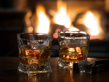 Téléchargez les photos : Deux verres de whisky et de glace sur une table en bois avec cheminée en arrière-plan - en image libre de droit