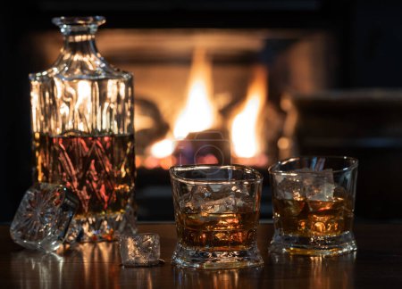 Photo for Two glasses of wiskey with ice and decanter on wooden table with burning fireplace in background - Royalty Free Image