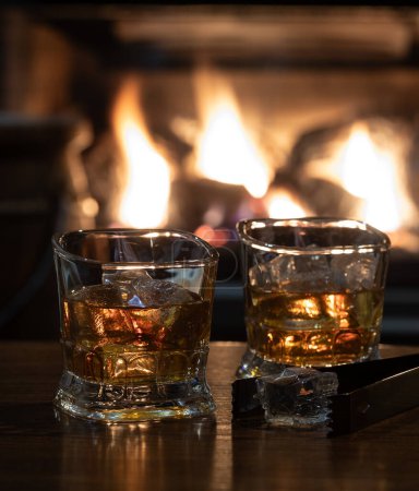 Photo for Two glasses of whiskey and ice on wooden table with burning fireplace in background - Royalty Free Image