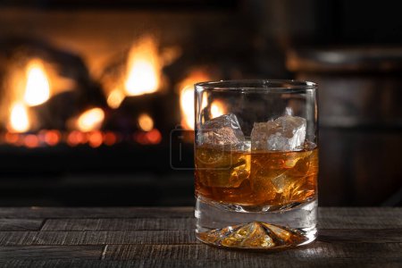 Photo for Glass of whiskey and ice on wooden table with burning fireplace in background - Royalty Free Image