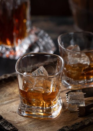 Téléchargez les photos : Deux verres de whisky avec glace sur plateau en bois rustique avec carafe dans le dos - en image libre de droit