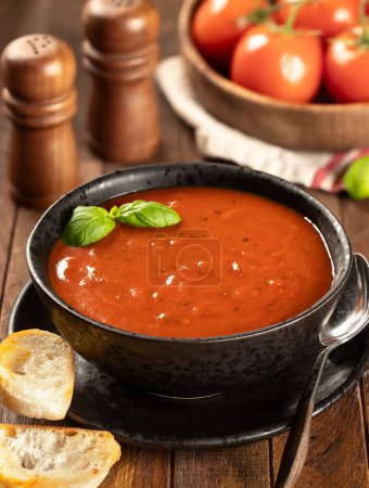 Photo for Bowl of tomato soup garnished with basil leaves and toasted baguette slices, fresh tomatoes in background on wooden table - Royalty Free Image