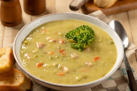 Photo for Bowl of split pea soup with ham and carrots garnished with parsley and toasted baguette on a wooden table - Royalty Free Image