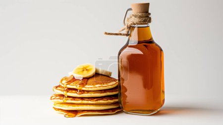A stack of pancakes drizzled with syrup beside a bottle of maple syrup on a white background 