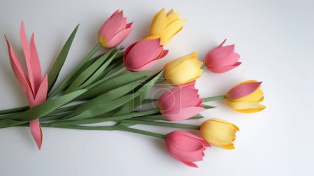 A collection of delicate paper tulips in shades of pink and yellow, artfully arranged with their green stems splayed across a bright white background.