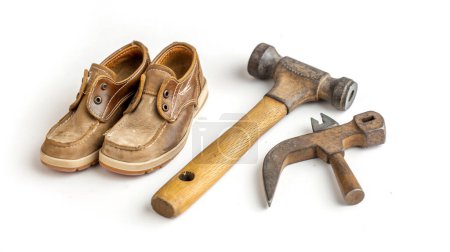 Paire de chaussures marron usées à côté d'un marteau vintage et d'un outil de cordonnier sur fond blanc, symbolisant l'artisanat.