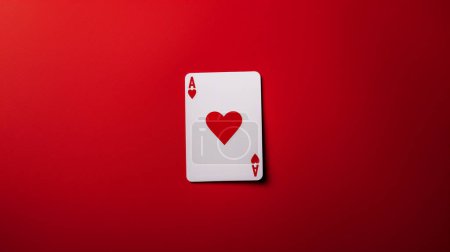 Photo for The Ace of Hearts playing card lies flat on a vibrant red background, creating a striking and bold visual contrast with its simple, iconic design. - Royalty Free Image