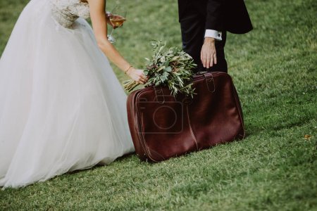 Téléchargez les photos : Mariée et marié dans le parc avec sac en cuir et bouquet de fleurs de champ - en image libre de droit