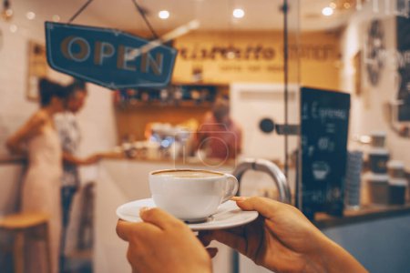 Foto de Coffee cup latte art in women's hands in background of interior of coffee shop. Local small businesses at food service. - Imagen libre de derechos