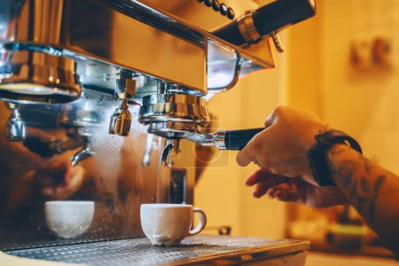 Foto de Professional coffee brewing in coffee shop. Close-up photo of espresso pouring from coffee machine. Barista make coffee cup in warm cozy atmosphere in small cafe. - Imagen libre de derechos