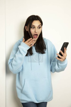 Photo for Emotional girl with a phone in a blue jacket on a light background. The model is shocked. - Royalty Free Image
