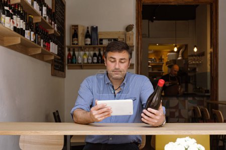 Foto de Manager sitting in his wine shop and wine bar checks the label of a bottle of wine using his tablet - Imagen libre de derechos