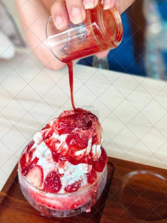 Strawberry bingsu in a glass cup on a wooden tray. Colorful, delicious fruit ice cream topped with syrup is ready to serve.