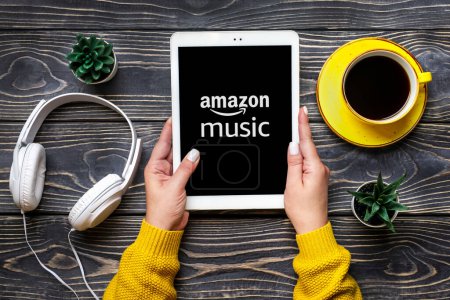 Photo for Bangkok, Thailand October 2022 Amazon music online music streaming platform and store, on screen of tablet in hand, wireless headphones, charger capsule on wooden table. - Royalty Free Image
