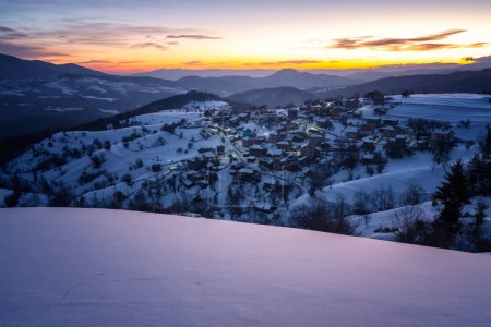 Téléchargez les photos : Belle vue sur le lever du soleil avec des pentes de montagne enneigées et petit village parmi eux dans la matinée gelée d'hiver, les montagnes Rhodopi, Bulgarie - en image libre de droit