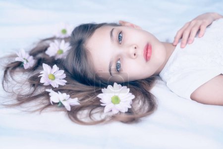 Foto de Primer plano retrato en tonos pastel de una hermosa chica caucásica acostada en una cama con flores de margarita en su pelo largo. - Imagen libre de derechos