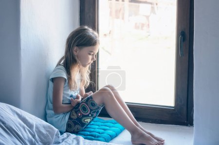 Photo for Vintage portrait of a beautiful little girl with bare feet and pensive gaze sitting near a window inside a room - Royalty Free Image