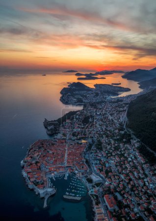 Atemberaubende Luftaufnahme der malerischen Stadt Dubrovnik mit der Altstadt, beleuchteten Straßen und Gebäuden und dem Yachthafen mit Booten bei feurigem Sonnenuntergang.