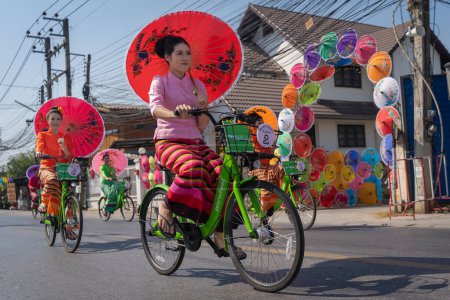 Téléchargez les photos : Chiang Mai, Thailand - January 20, 2023: Pretty women holding beautiful umbrellas in traditional costumes and riding cycling bicycles annual show at San Kamphaeng, Bosang umbrella festival. - en image libre de droit