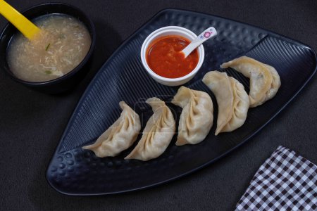 Photo for Picture of steamed chicken stuffed momo served in a black plate along with chicken soup in a bowl and sauce on black background. - Royalty Free Image
