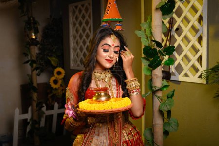 Téléchargez les photos : Portrait de jolie jeune fille indienne portant saree traditionnelle, bijoux en or et bracelets tenant plaque d'offrande religieuse dans l'éclairage d'atelier intérieur. Culture, religion et mode indiennes. - en image libre de droit