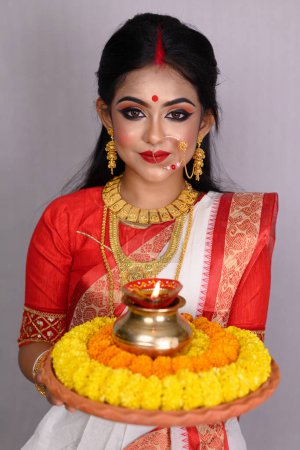 Photo for Portrait young Indian girl wearing traditional saree, gold jewellery, bangles holding plate of religious offering and standing in front of white background. Indian culture, religion and festivals. - Royalty Free Image