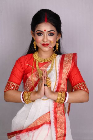 Foto de Retrato de una hermosa joven india vestida con un saree indio tradicional, joyas de oro y brazaletes de pie frente a un fondo blanco. Maa Durga agomoni disparar concepto. - Imagen libre de derechos