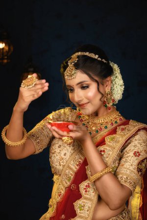 Portrait of a pretty young Indian woman dressed in traditional lehenga, gold jewellery and bangles holding diya in hands on decorative background. Indian culture, occasion, religion and fashion.