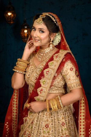 Photo for Stunning Indian bride dressed in traditional bridal lehenga with heavy gold jewellery and veil smiles tenderly in studio lighting. Wedding fashion and lifestyle. - Royalty Free Image