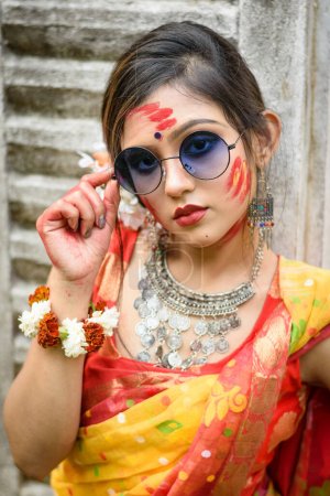 Porträt eines hübschen jungen Mädchens, das traditionellen indischen Sari und Schmuck trägt, mit Farben spielt und modisch mit Sonnenbrille auf dem Fest der Farben namens Holi posiert.