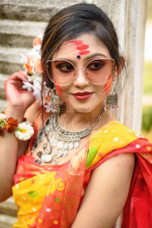 Porträt eines hübschen jungen Mädchens, das traditionellen indischen Sari und Schmuck trägt, mit Farben spielt und modisch mit Sonnenbrille auf dem Fest der Farben namens Holi posiert.