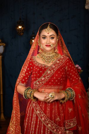 Stunning Indian bride dressed in traditional red bridal lehenga with heavy gold jewellery and veil smiles tenderly in studio lighting. Wedding fashion and lifestyle.