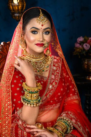 Atemberaubende indische Braut in traditionellen roten Braut Lehenga mit schwerem Goldschmuck und Schleier sitzt in einem Stuhl lächelt zärtlich in Studio-Beleuchtung gekleidet. Hochzeitsmode und Lifestyle.