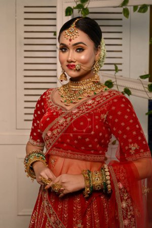 Impresionante novia india adornada con una lehenga tradicional de novia roja con gracia lleva joyas de oro pesado y un velo y sonríe tiernamente en la iluminación del estudio. Boda moda y estilo de vida.
