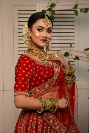 Stunning Indian bride adorned in a traditional red bridal lehenga gracefully wears heavy gold jewelry and a veil and smiles tenderly in studio lighting. Wedding fashion and lifestyle.