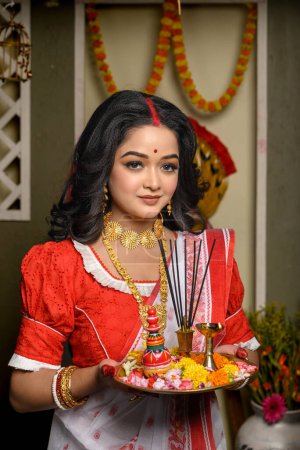 Portrait of a young woman, adorned in a traditional saree, complemented by gold jewellery and bangles, holding a plate filled with religious offerings. Indian culture, religion and festivals.
