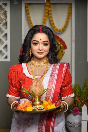 Porträt einer jungen Frau, geschmückt mit einem traditionellen Sari, ergänzt durch Goldschmuck und Armreifen, die einen Teller mit religiösen Opfergaben in der Hand hält. Indische Kultur, Religion und Feste.