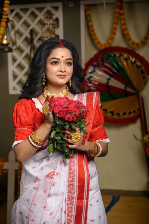 Porträt einer eleganten jungen Frau in einem traditionellen Sari, verziert mit Goldschmuck und Armreifen, die zart Blumen in ihren Händen hält. Indische Kultur, Religion und Mode.