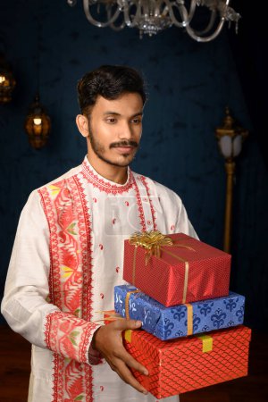 Porträt eines jungen, gut aussehenden Mannes, der in einen traditionellen Kurta-Pyjama gekleidet ist und unter Studioleuchtung Geschenkschachteln in den Händen hält. Indische Mode, Veranstaltungen und Lifestyle.