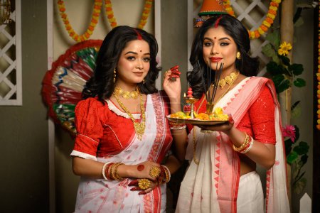 Mujeres indias, adornadas con saris tradicionales y joyas de oro, incluyendo brazaletes, sosteniendo un plato lleno de ofrendas religiosas. Festival indio, cultura, ocasión, religión y moda étnica.