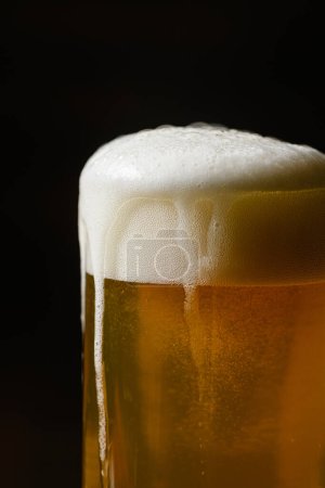 Kaltes Bier mit Schaum im Becher vor dunklem Hintergrund, Nahaufnahme.