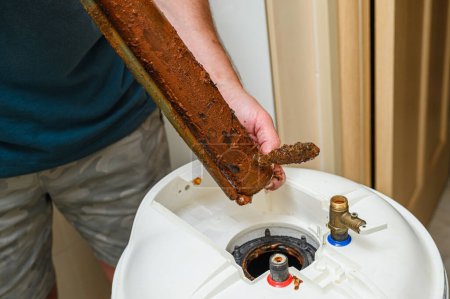 El hombre sostiene en su mano un elemento calefactor eléctrico dañado. Reparación de la caldera, sustitución del elemento calefactor de agua roto.
