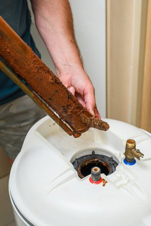 El hombre sostiene en su mano un elemento calefactor eléctrico dañado. Reparación de la caldera, sustitución del elemento calefactor de agua roto.