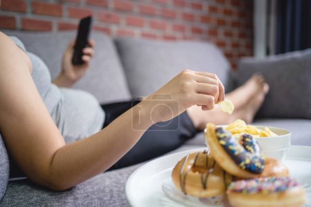 Foto de Mujer gorda acostada en el sofá sosteniendo el teléfono inteligente y comiendo papas fritas, concepto de comer en exceso - Imagen libre de derechos
